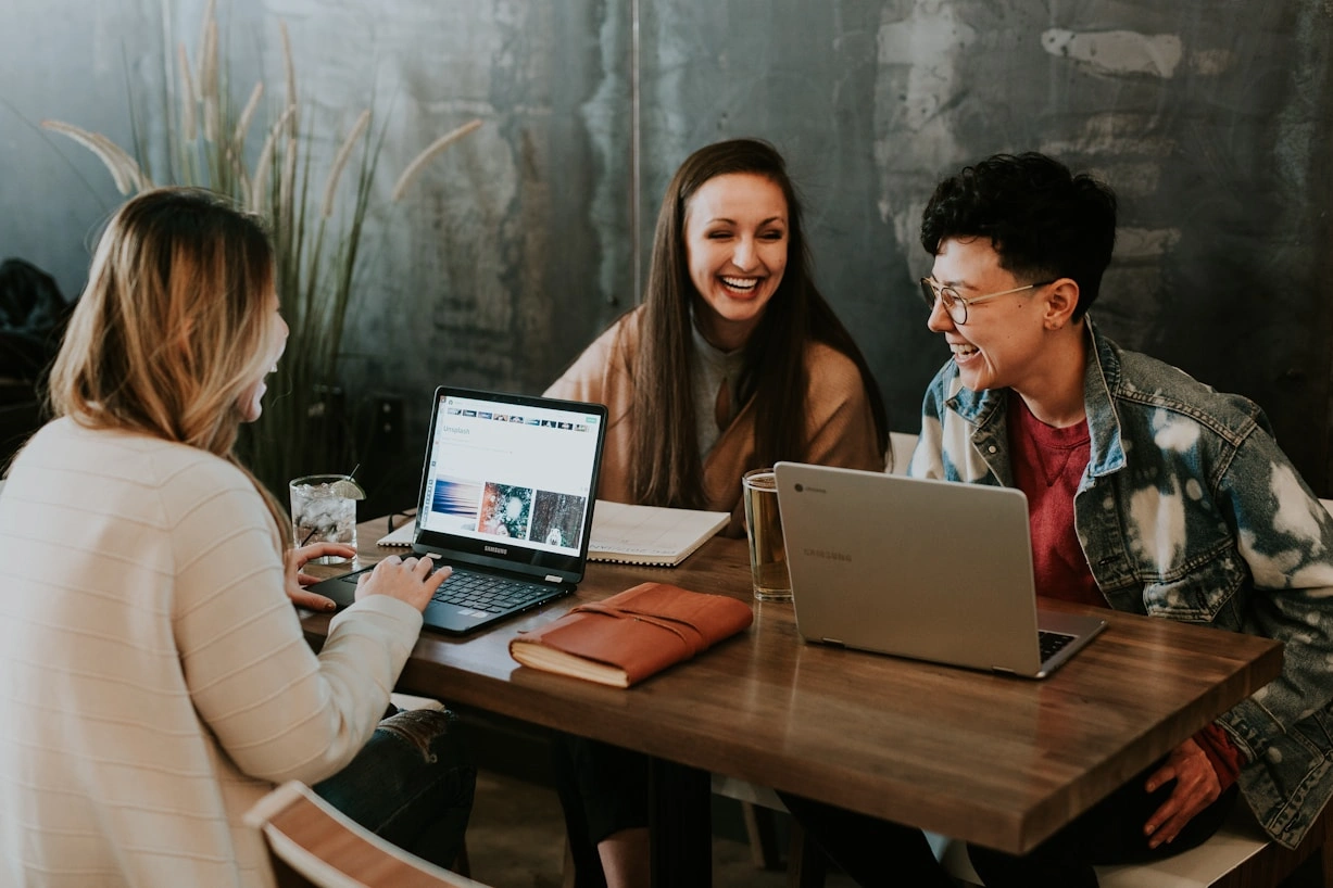 Three colleagues laughing together over laptops at a shared table